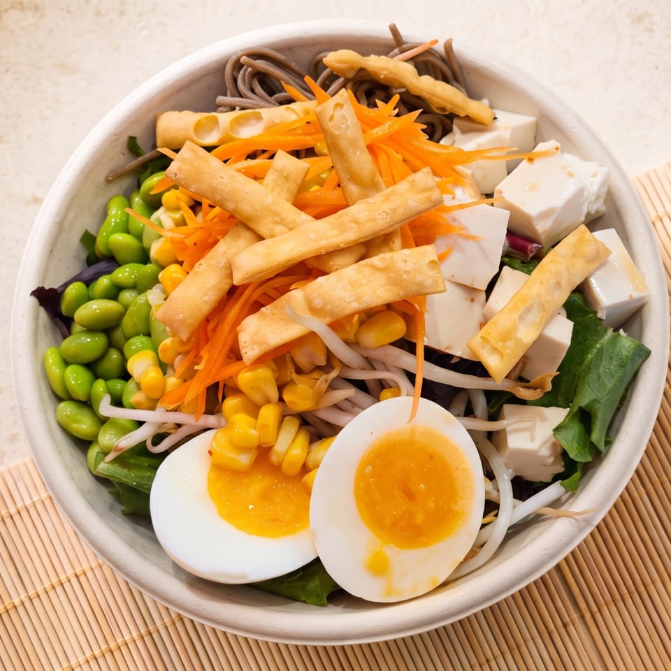 A colorful Asian-inspired salad bowl with noodles, edamame, corn, tofu, carrots, and a soft-boiled egg.