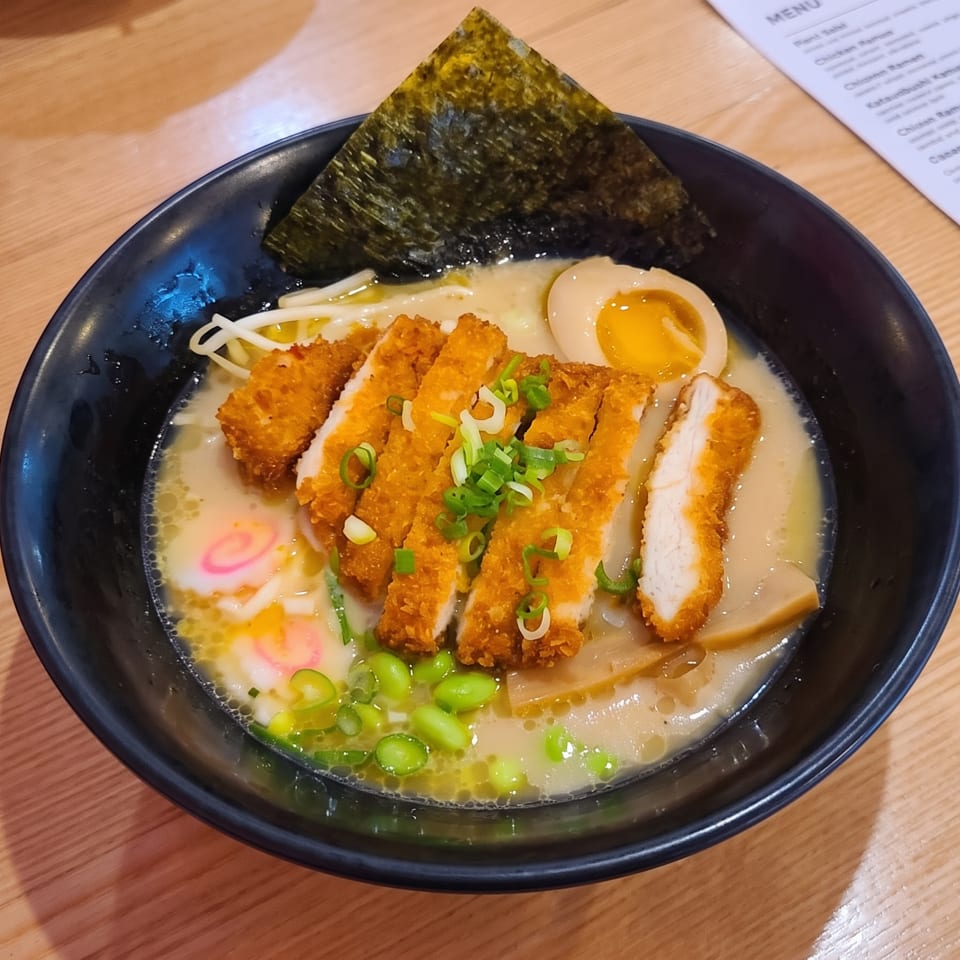 A bowl of ramen with crispy chicken katsu, a soft-boiled egg, nori, and green onions.