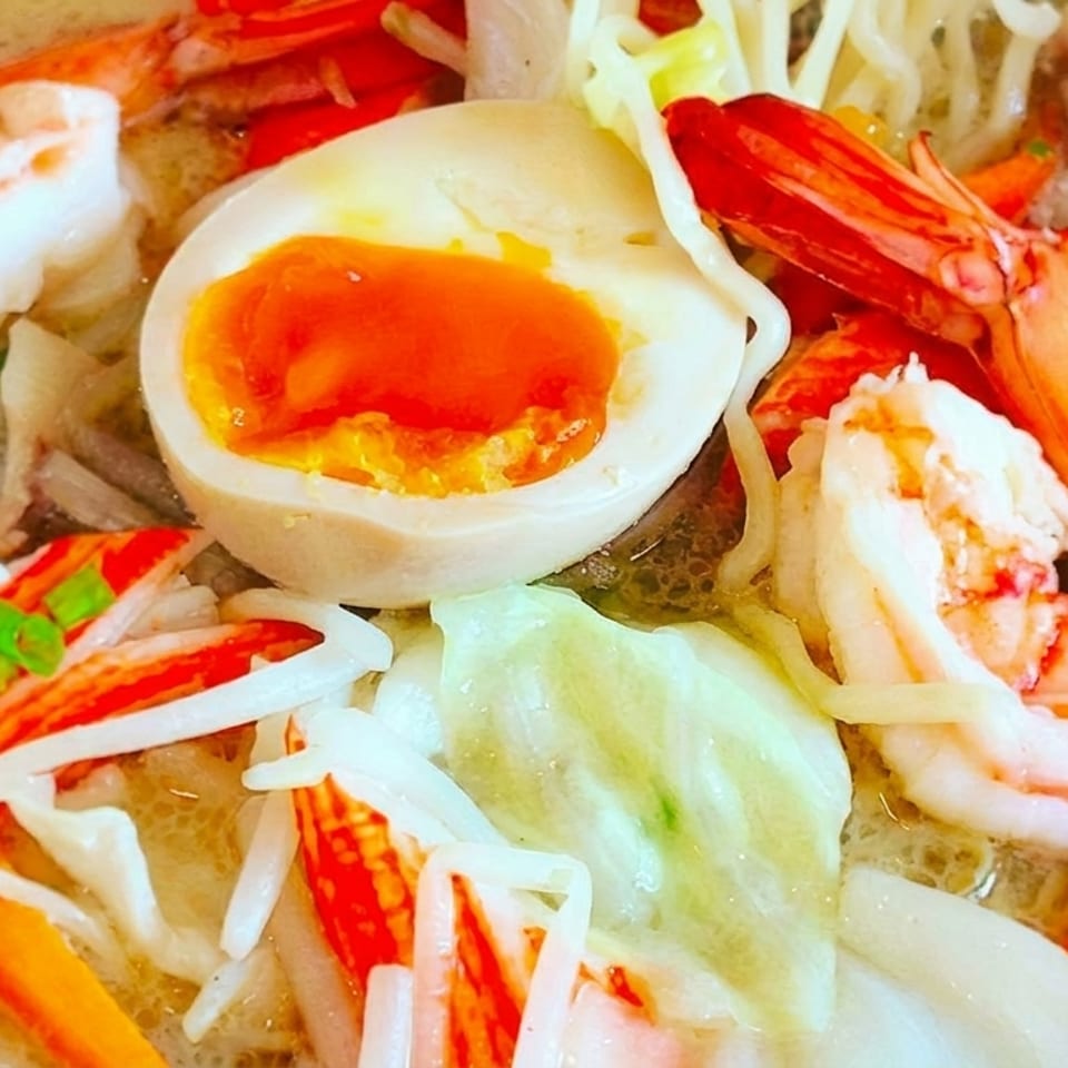 Close-up of a ramen bowl with shrimp, crab sticks, soft-boiled egg, and vegetables.