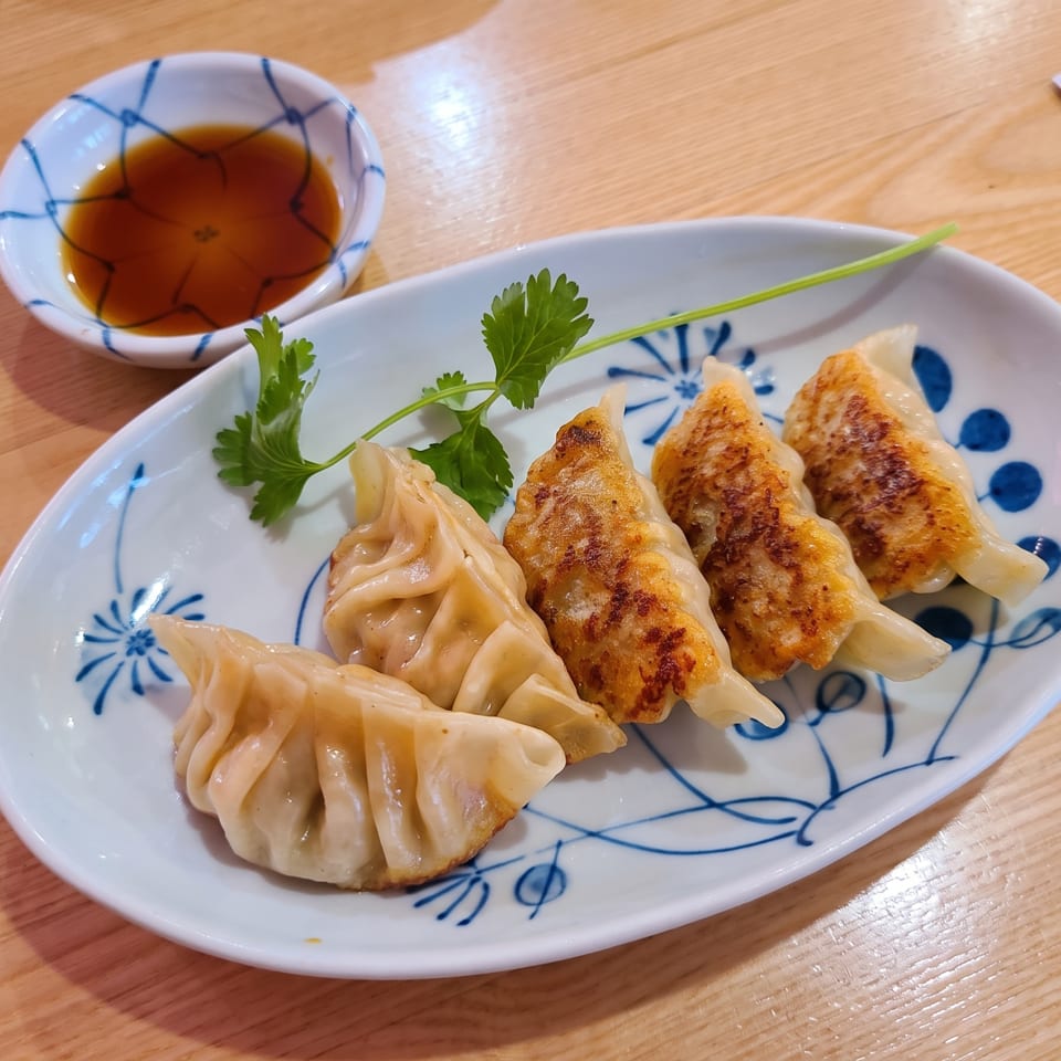 Five pan-fried gyoza dumplings with cilantro on a white plate, with a small bowl of soy sauce.