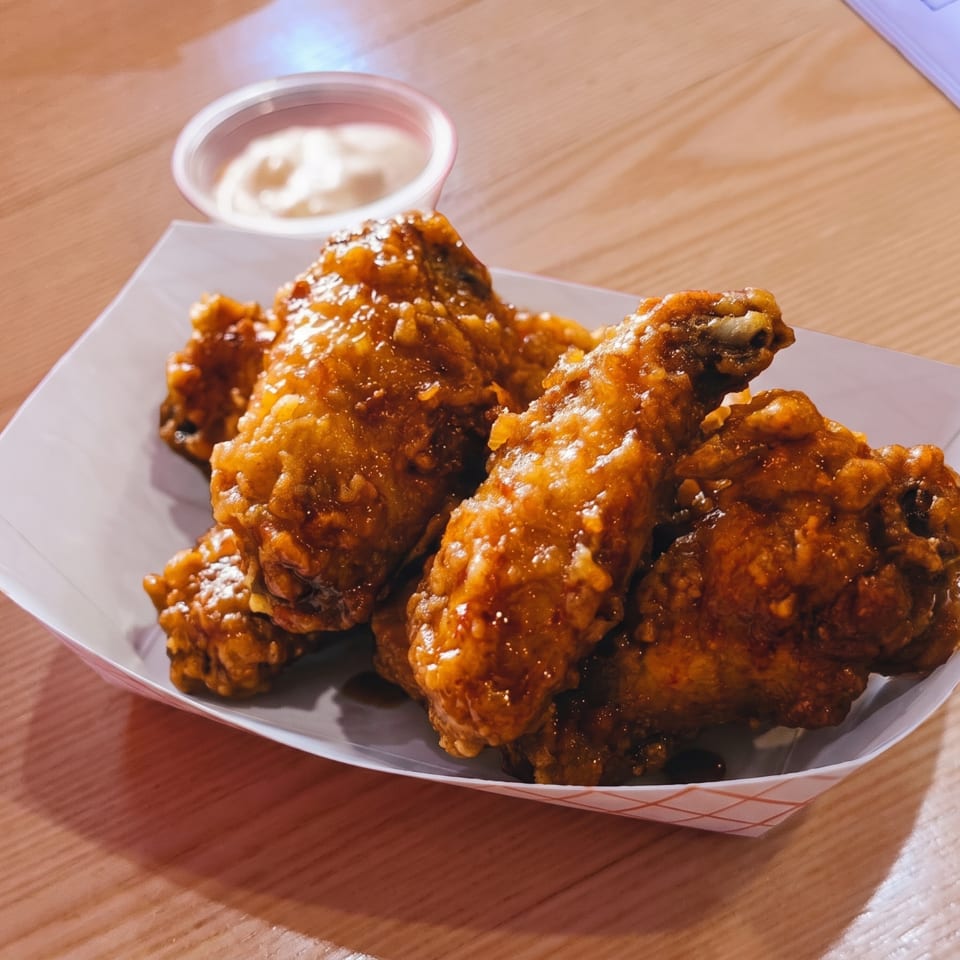 Crispy, glazed chicken wings in a paper tray with a side of dipping sauce on a wooden table.